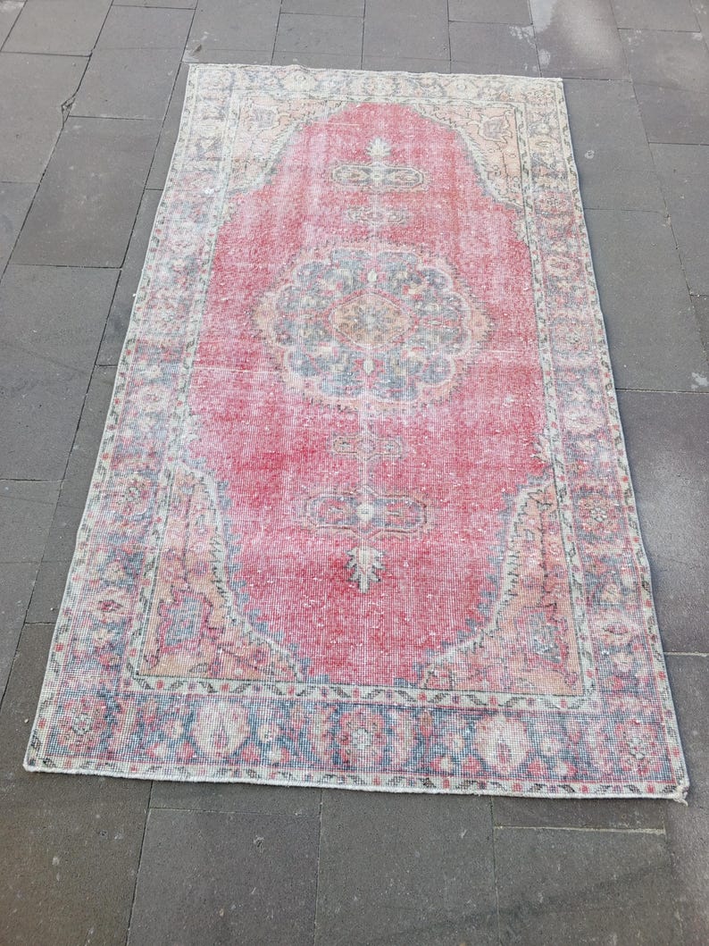 May include: A rectangular, distressed rug with a faded red central field and a detailed border. The rug features a central medallion design and intricate patterns in shades of blue, green, and cream. The rug's worn appearance suggests it is vintage.