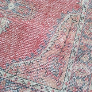 May include: A close-up view of a distressed, vintage rug with a faded red central field. The rug features intricate border designs in shades of blue, beige, and peach. The rug's texture is visible, showcasing its aged appearance.