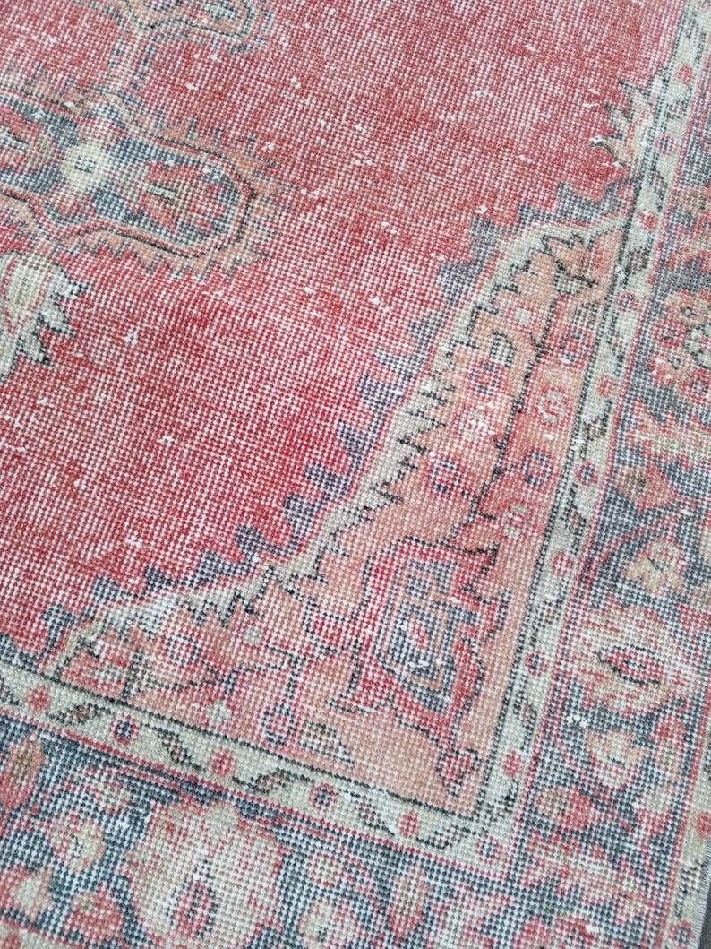 May include: A close-up view of a distressed, vintage rug with a faded red central field. The rug features intricate border designs in shades of blue, beige, and peach. The rug's texture is visible, showcasing its aged appearance.