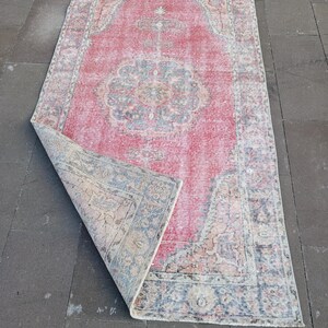 May include: A rectangular, distressed rug with a faded red central field and a detailed border. The rug features a central medallion design in shades of blue, beige, and pink. The corner of the rug is folded over, revealing the underside.