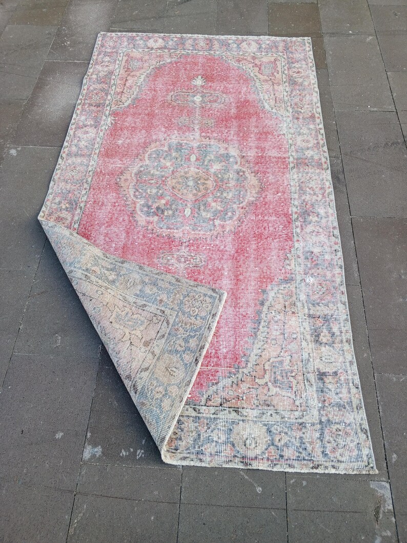 May include: A rectangular, distressed rug with a faded red central field and a detailed border. The rug features a central medallion design in shades of blue, beige, and pink. The corner of the rug is folded over, revealing the underside.