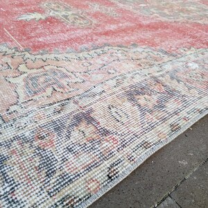 May include: A distressed, rectangular rug with a faded red central field and intricate border details. The rug features a traditional pattern with muted tones of beige, brown, and blue, creating a vintage aesthetic. The rug's edges are frayed, adding to its aged appearance.
