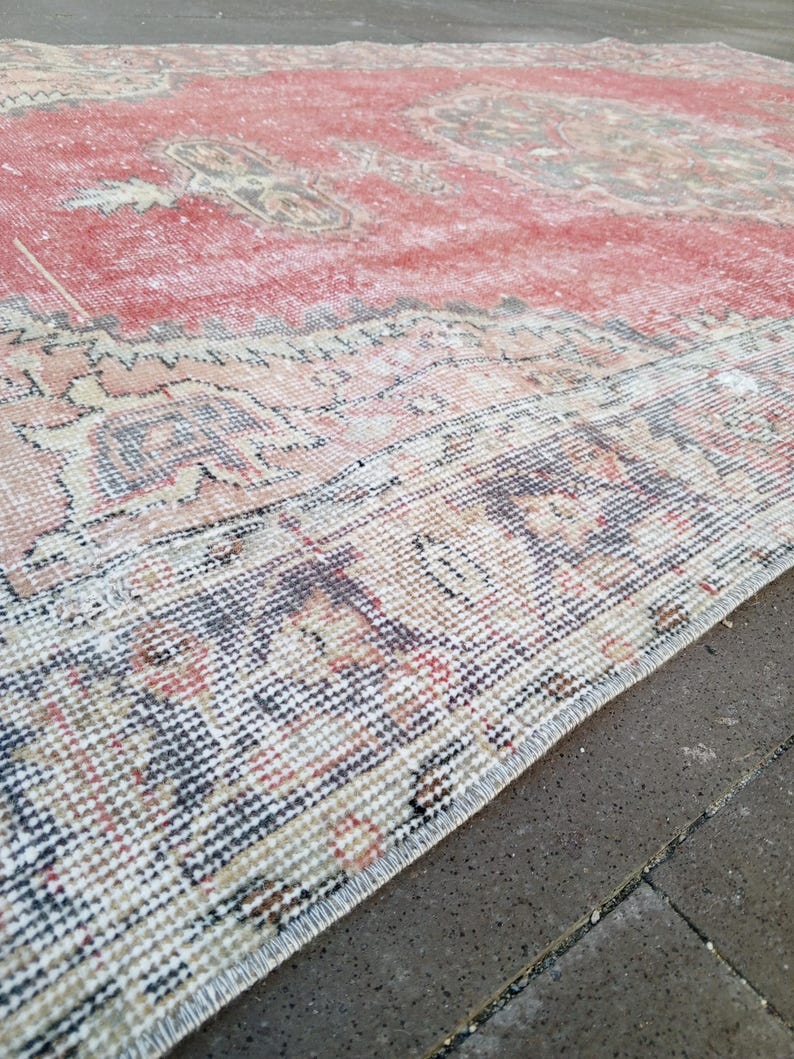 May include: A distressed, rectangular rug with a faded red central field and intricate border details. The rug features a traditional pattern with muted tones of beige, brown, and blue, creating a vintage aesthetic. The rug's edges are frayed, adding to its aged appearance.