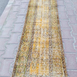 May include: A long, narrow rug with a faded, vintage look. The rug has a brown and yellow color scheme with a subtle pattern. The rug is made of a woven material and has a fringed edge.
