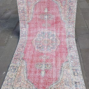 May include: A rectangular, distressed rug with a central red field and intricate border designs. The rug features faded patterns in shades of blue, beige, and pink, creating a vintage aesthetic. The rug is held up, showcasing its full length.