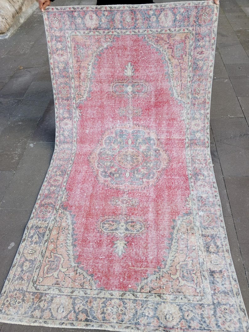 May include: A rectangular, distressed rug with a central red field and intricate border designs. The rug features faded patterns in shades of blue, beige, and pink, creating a vintage aesthetic. The rug is held up, showcasing its full length.
