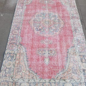 May include: A rectangular, distressed rug with a central red field and intricate border details. The rug features a faded, vintage aesthetic with floral and geometric patterns in shades of blue, beige, and pink. The rug's dimensions are not visible.