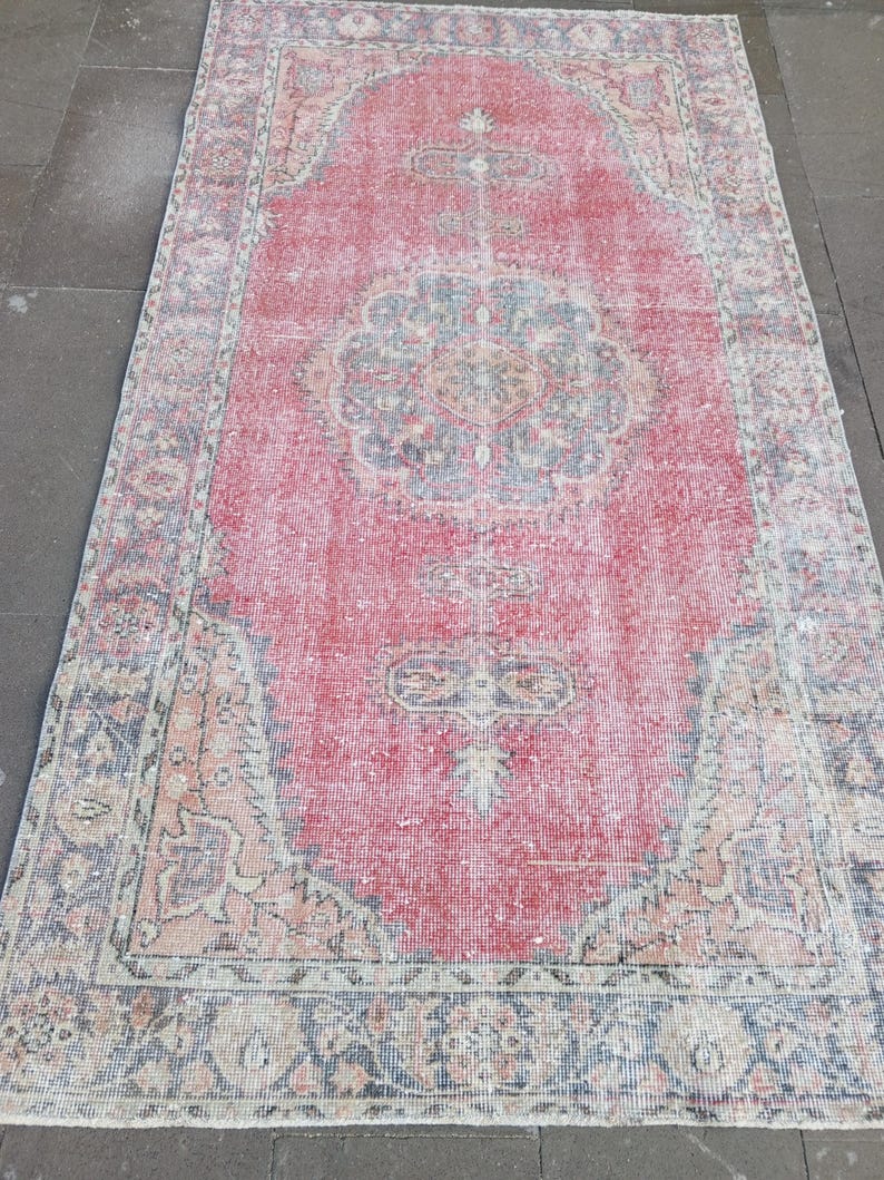 May include: A rectangular, distressed rug with a central red field and intricate border details. The rug features a faded, vintage aesthetic with floral and geometric patterns in shades of blue, beige, and pink. The rug's dimensions are not visible.