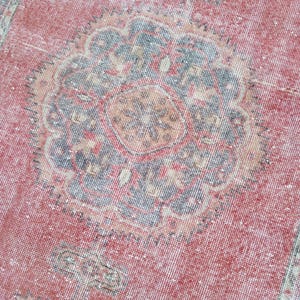 May include: Close-up of a distressed, vintage rug with a faded red background. The rug features a central medallion design in shades of blue, beige, and orange. The rug's texture is visible, with a worn appearance.
