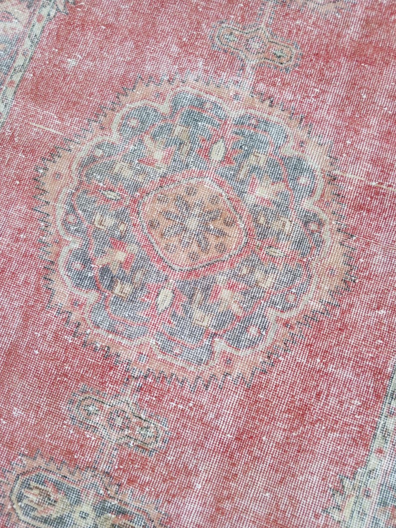 May include: Close-up of a distressed, vintage rug with a faded red background. The rug features a central medallion design in shades of blue, beige, and orange. The rug's texture is visible, with a worn appearance.