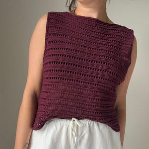 May include: A sleeveless, claret-coloured crochet top with a boat neck. The top features a horizontal openwork pattern. The model is wearing white drawstring trousers. The top is handmade and suitable for warm weather.