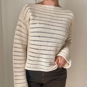 Op de afbeelding: Crèmekleurige gehaakte trui met lange mouwen en een boothals. De trui heeft een open breipatroon en een losse pasvorm. De mouwen zijn licht uitlopend aan de polsen. De trui wordt gecombineerd met een bruine broek.