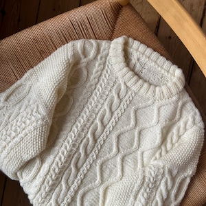 Könnte beinhalten: Cremefarbener Zopfstrickpullover mit aufwendigen Mustern. Der Pullover hat einen Rundhalsausschnitt und verschiedene Zopfmuster auf Körper und Ärmeln. Das Kleidungsstück wird auf einem Webstuhl präsentiert.