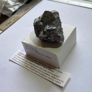 Può includere: Un campione minerale di Skutterudite grigio scuro dalla miniera di Bou Azzer, Marocco, appoggiato su una scatola bianca. Il minerale ha una lucentezza metallica. Un'etichetta fornisce dettagli sul campione e sul suo collezionista.
