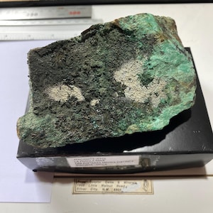 May include: A rough-hewn mineral specimen, primarily green and dark gray, with areas of white and brown. The mineral is displayed on a black base, with a label visible that includes the name "Atacamite" and the mine location in Chile.