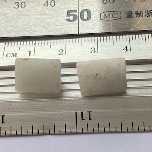 Op de afbeelding: Twee rechthoekige, crèmekleurige stenen met subtiele bruine markeringen worden getoond op een metalen liniaal. De liniaal toont centimeter metingen. De stenen zijn ongeveer 2,5 cm lang.