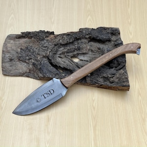May include: A handmade knife featuring a dark wooden handle and a broad, silver-coloured blade. The blade is engraved with the letters "TSD". The knife is presented on a piece of dark, textured wood.