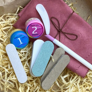 May include: A set of nail care tools in a pink pouch. The set includes two small jars of nail powder, two nail files, a cuticle pusher, and a small white spatula. The jars are labelled "Magic Girl" and have numbers "1" and "2" on them. The jars are labelled "Wax - Paste" and "Powder".