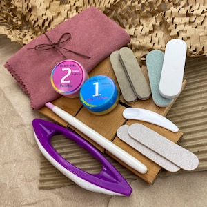 Puede incluir: Un conjunto de herramientas y productos para el cuidado de las uñas, que incluye un buffer de uñas morado, una lima de uñas blanca, una tabla de madera con limas de uñas, un pequeño frasco rosa etiquetado como "Magic Girl Cooling Coating 2 Powder", un pequeño frasco azul etiquetado como "Magic Girl Cooling Coating 1 Wax Paste" y un paño de microfibra rosa.
