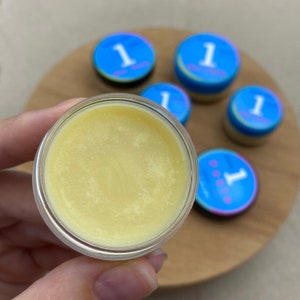 May include: A close-up of a small, clear jar filled with a pale yellow balm. The jar is being held by a hand. There are five other jars in the background, all with blue and white labels that say "1".