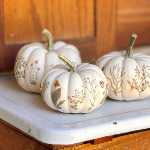 Puede incluir: Tres calabazas blancas decoradas con flores y hojas prensadas. Las calabazas están dispuestas sobre una superficie blanca con un fondo de madera.