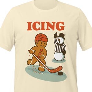 May include: Cream-colored t-shirt featuring a gingerbread man playing hockey with a snowman referee. The word "ICING" is printed above the cartoon characters. The gingerbread man wears a helmet and holds a hockey stick. The snowman is dressed as a referee.