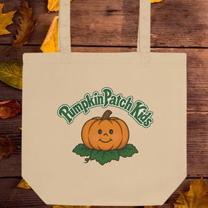 May include: Beige tote bag with the text "Pumpkin Patch Kids" in green script above a smiling orange pumpkin on green leaves. The bag has two beige handles and is set against a wooden background with autumn leaves.