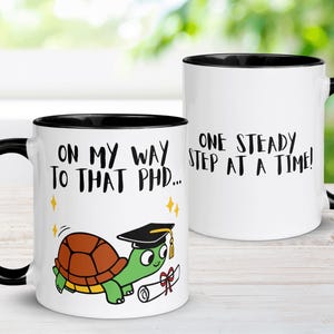 May include: White ceramic mug with a black rim. The mug has a cartoon turtle wearing a graduation cap and holding a diploma. The text on the mug reads "On my way to that PhD... One steady step at a time!"