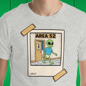 May include: A gray t-shirt with a graphic of a green alien walking into a bathroom labeled "Area 52". The graphic is framed with a white border and has a yellow tape sticker on the top left corner. The text "1947" is printed below the graphic.