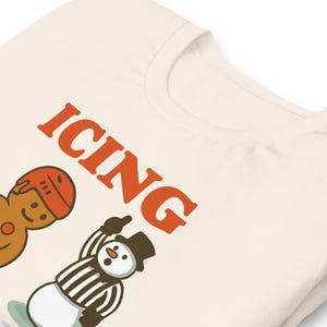 May include: Cream-colored t-shirt with the word "ICING" in orange block letters. The shirt features cartoon illustrations of a gingerbread man wearing a hockey helmet and a snowman referee. The snowman is pointing upwards.