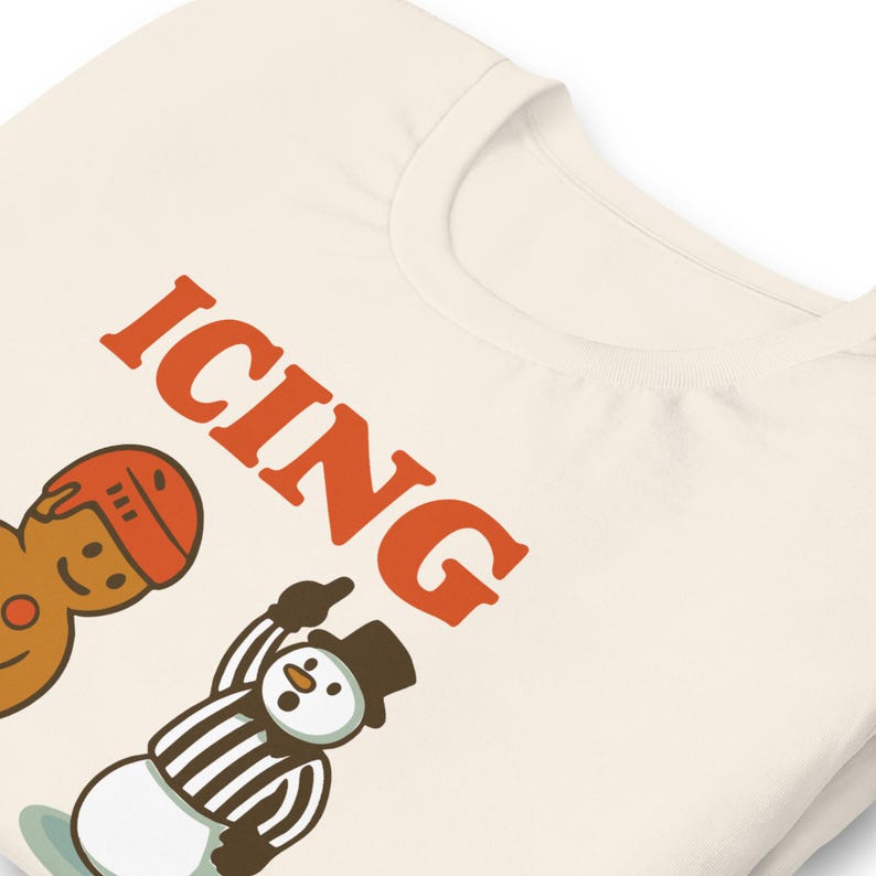 May include: Cream-colored t-shirt with the word "ICING" in orange block letters. The shirt features cartoon illustrations of a gingerbread man wearing a hockey helmet and a snowman referee. The snowman is pointing upwards.