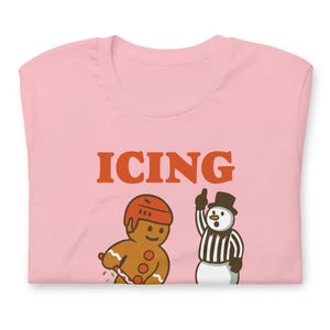 May include: A light pink t-shirt with the word "ICING" in orange block letters. Below the text are cartoon illustrations of a gingerbread man in a hockey helmet and a snowman referee.