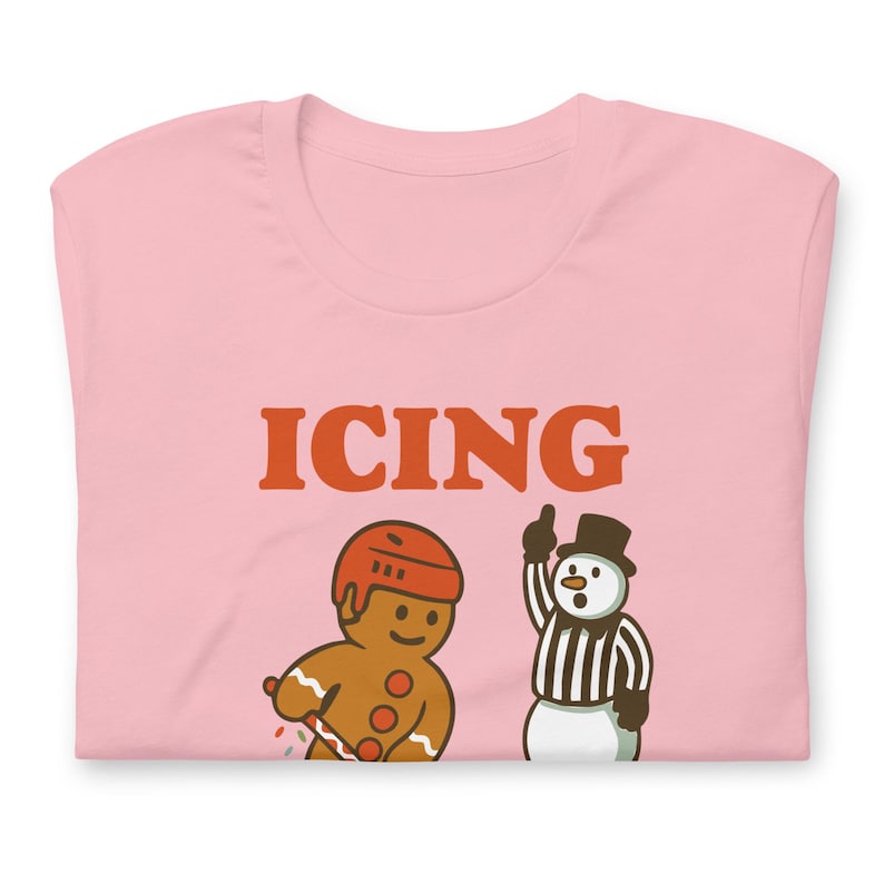 May include: A light pink t-shirt with the word "ICING" in orange block letters. Below the text are cartoon illustrations of a gingerbread man in a hockey helmet and a snowman referee.