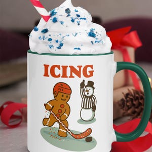 May include: White ceramic mug with a green handle and rim, featuring the word "ICING" in orange. The mug has a gingerbread man playing hockey and a snowman referee. Topped with whipped cream and a red and white striped straw.