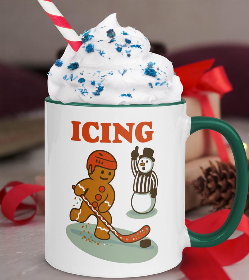 May include: White ceramic mug with a green handle and rim, featuring the word "ICING" in orange. The mug has a gingerbread man playing hockey and a snowman referee. Topped with whipped cream and a red and white striped straw.