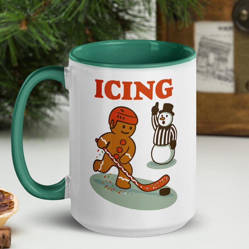 May include: White ceramic mug with a green handle and interior. The mug features a gingerbread man playing hockey with a snowman referee, and the word "ICING" in red. A fun, festive design.