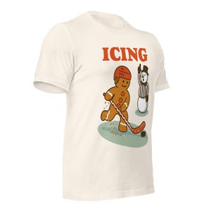 May include: Off-white t-shirt with a cartoon gingerbread man playing hockey, wearing a red helmet. The word "ICING" is printed in red above the gingerbread man. A snowman referee is nearby. The shirt is a fun, novelty item.