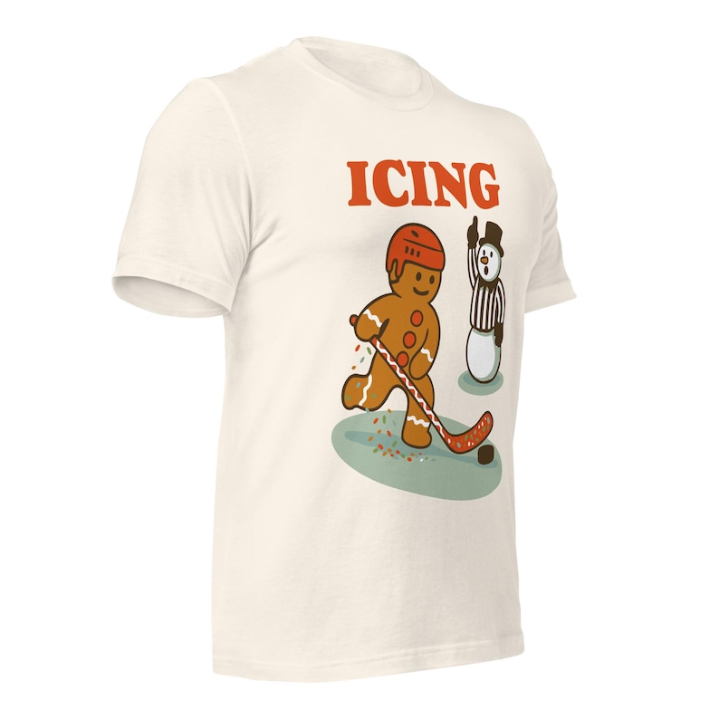 May include: Off-white t-shirt with a cartoon gingerbread man playing hockey, wearing a red helmet. The word "ICING" is printed in red above the gingerbread man. A snowman referee is nearby. The shirt is a fun, novelty item.