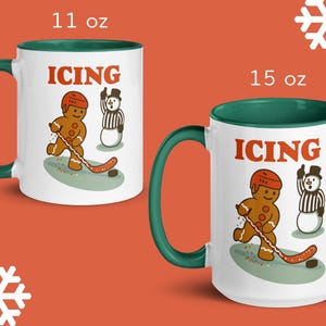 May include: Two white ceramic mugs with green handles and rims. The mugs feature a gingerbread man playing hockey with the word "ICING" in orange. One mug is 11 oz, the other is 15 oz.