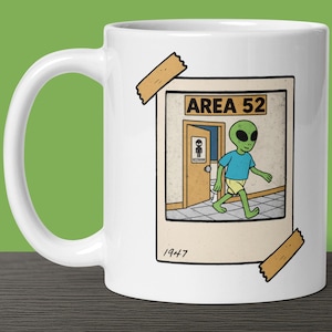 May include: White ceramic mug with a graphic of an alien entering a restroom labeled "Area 52". The image is framed with a brown border and has a vintage look. The text "1947" is printed below the image.
