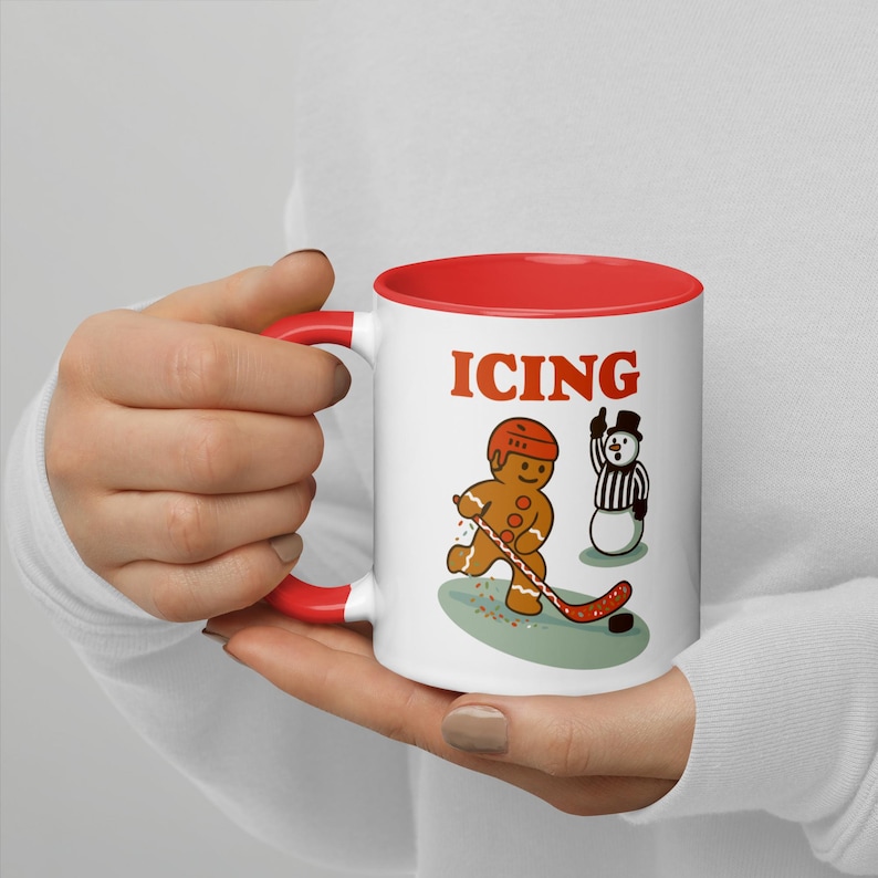 May include: White ceramic mug with a red interior and handle. The mug features a cartoon gingerbread man playing hockey with the word "ICING" above the image. A snowman referee is also depicted.