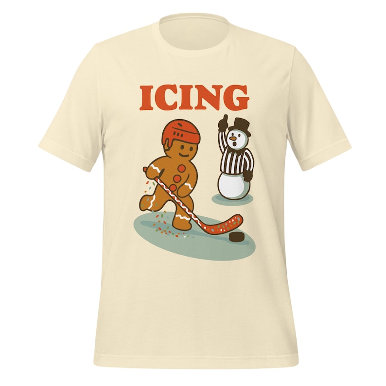 May include: Cream-colored t-shirt featuring a gingerbread man playing hockey with a snowman referee. The word "ICING" is printed above the cartoon characters. The gingerbread man wears a helmet and holds a hockey stick. The snowman is dressed as a referee.
