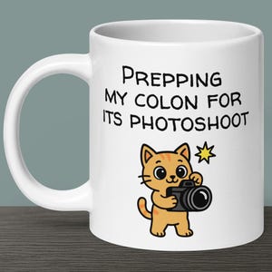 May include: White ceramic mug featuring a black and white illustration of a cat holding a camera. The text on the mug reads "Prepping my colon for its photoshoot."