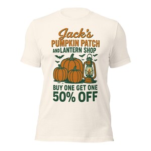 Puede incluir: Camiseta color crema con un diseño de "Jack's Pumpkin Patch and Lantern Shop". El diseño incluye calabazas naranjas, una linterna y murciélagos, con el texto "Buy One Get One 50% Off".