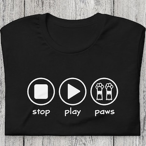 May include: Black t-shirt with a white graphic. The design includes a stop button, a play button, and two paw prints, each within a circle. Underneath are the words "stop", "play", and "paws".