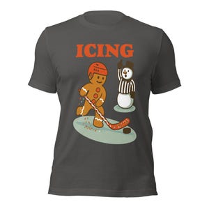 May include: Dark gray t-shirt with the word "ICING" in orange block letters. The shirt features a gingerbread man playing hockey, with a snowman referee. The gingerbread man is wearing a helmet and the snowman is raising his arm.