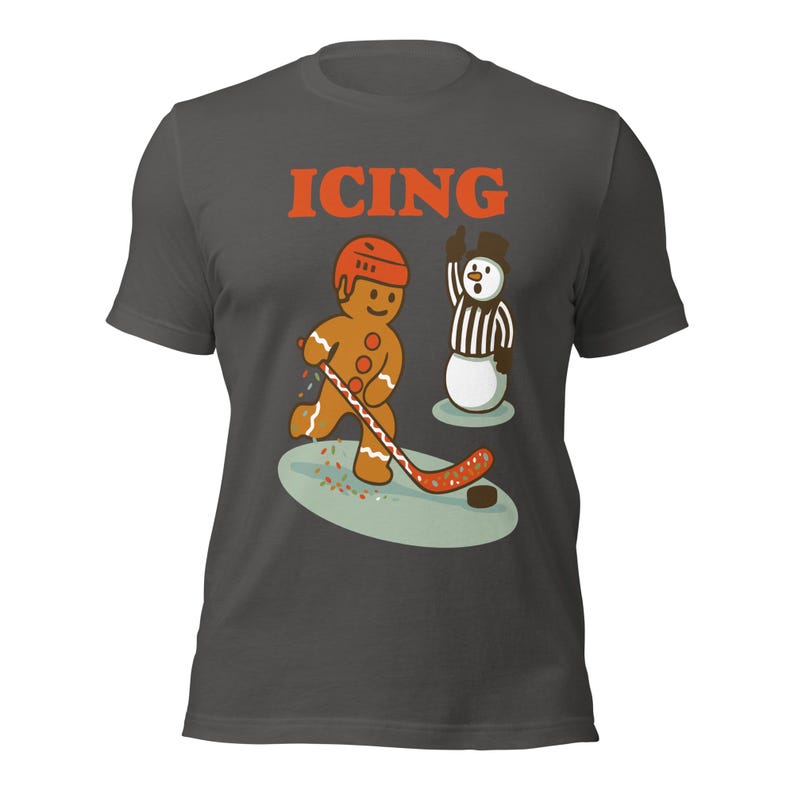 May include: Dark gray t-shirt with the word "ICING" in orange block letters. The shirt features a gingerbread man playing hockey, with a snowman referee. The gingerbread man is wearing a helmet and the snowman is raising his arm.