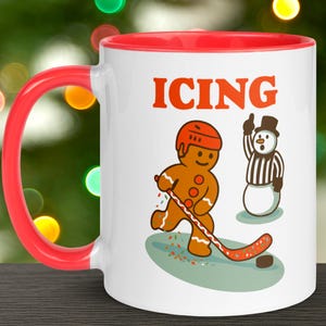May include: White ceramic mug with a red handle and interior. The mug features a cartoon gingerbread man playing hockey with the word "ICING" in orange above the image. A snowman referee is also depicted.