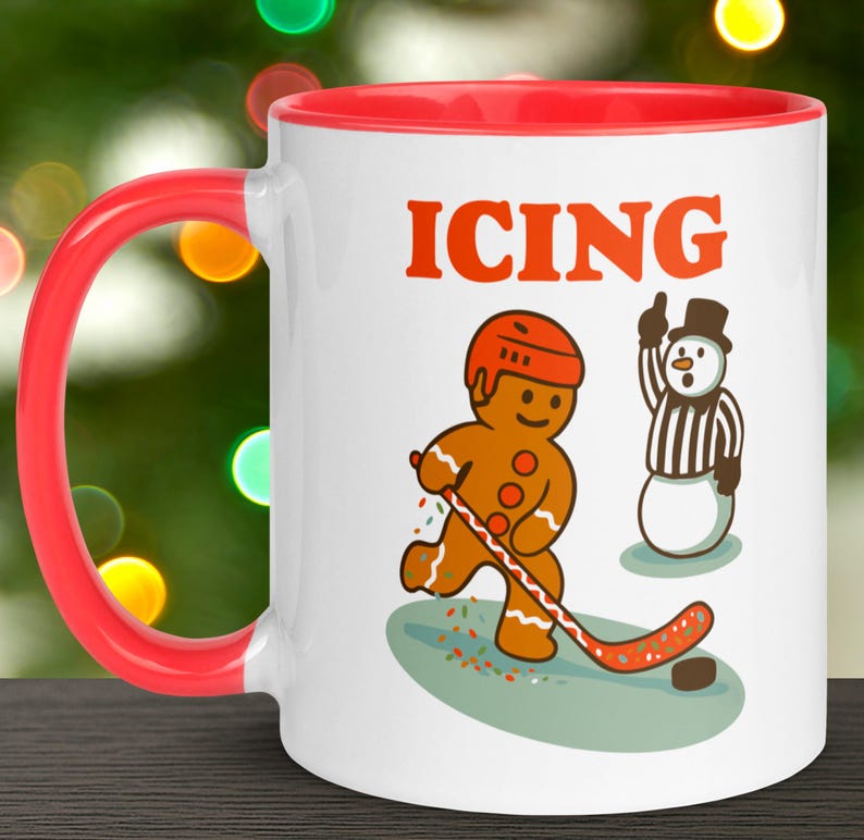 May include: White ceramic mug with a red handle and interior. The mug features a cartoon gingerbread man playing hockey with the word "ICING" in orange above the image. A snowman referee is also depicted.