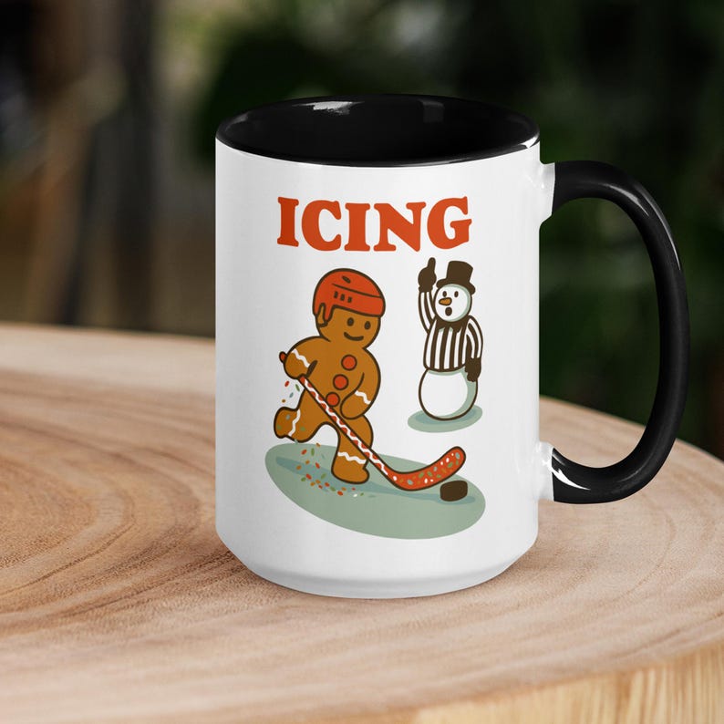May include: White ceramic mug with a black interior and handle. The mug features a cartoon gingerbread man playing hockey with a snowman referee. The word "ICING" is printed above the characters in orange.